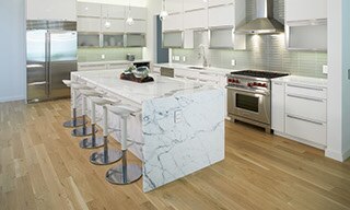 Modern kitchen with white & gray waterfall island, white & silver bar stools, wood look floor tiling, white quartz countertops, light green glass backsplash, and white cabinets.
