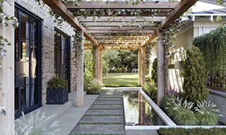 Outdoor courtyard with gray 2CM stone look porcelain pavers, coy pond surrounded by fountain grass, covered by a natural wood pergola with flowering vines.