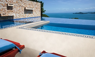 Infinity pool with white tile deck with blue accents, red lounge chairs with blue cushions, and an ocean view with blue skies in the background.