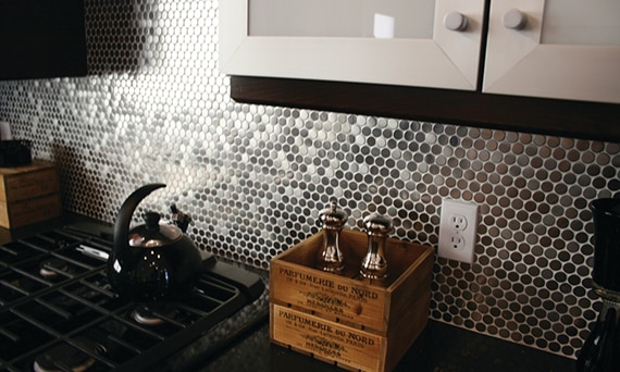 Black gas stove, black tea pot, stainless steel penny round mosaic backsplash, black countertop, and décor crate holding metal pepper grinder & salt shaker.