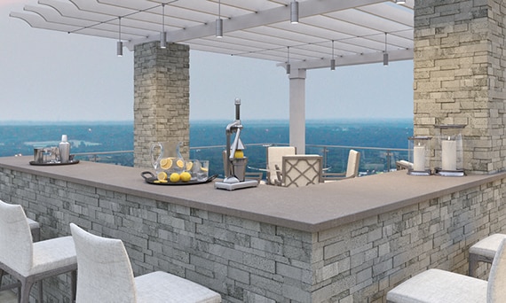 Rooftop patio with stacked stone-faced bar under a pergola, gray quartz countertops, pitcher of lemon water, and gray linen-covered bar stools.