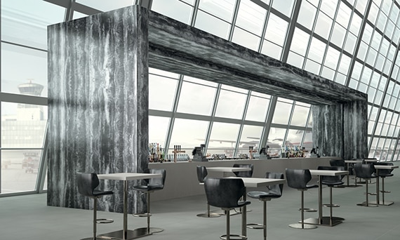 Luxury airport bar with walls of glass and tall ceilings. Bar surrounded by a canopy like architecture covered in black marble-look porcelain slab. Floors feature cool gray tile.