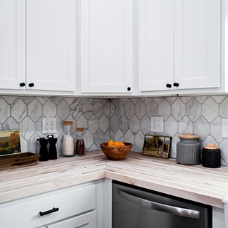 Kitchen backsplash of white & gray, Victorian mosaic marble tile, butcher block countertop, white cabinets, and stainless steel dishwasher.