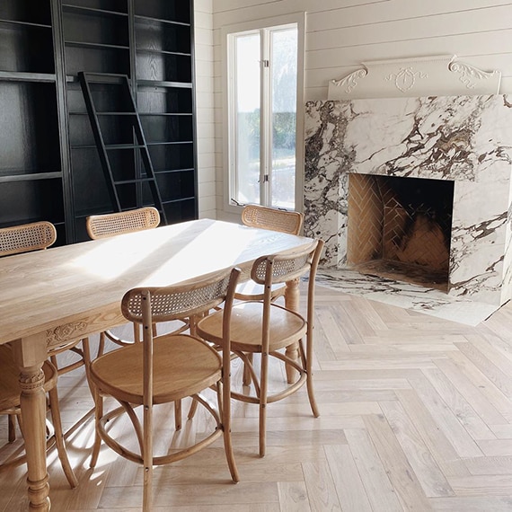 Newly renovated living room with herringbone wood flooring, white & brown fireplace surround that looks like marble, black bookcase, wood table & chairs.