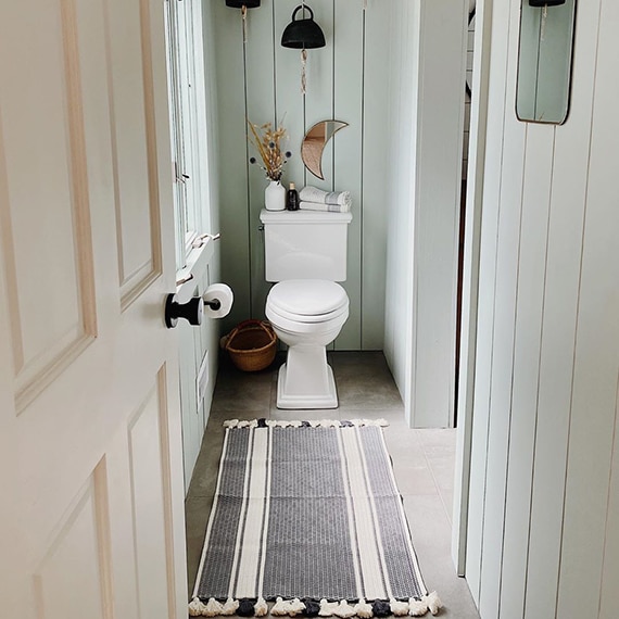 Newly renovated bathroom with gray floor tile that looks like concrete, vertical white shiplap walls, black matte fixtures, and black & wall tasseled rug.