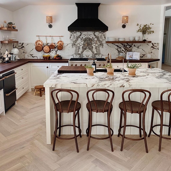Newly renovated kitchen with white & brown backsplash and countertops that look like marble, black vent hood, white kitchen island with sink and wicker stools.