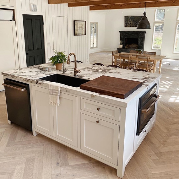 Newly renovated kitchen/family room with white kitchen island with sink and white & brown countertop that looks like marble, black fireplace, wood table & chairs.