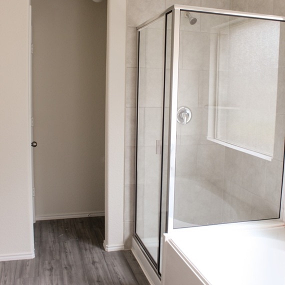 Before photo of builder-grade shower with beige tile, silver aluminum frame, frosted window, and separate bathtub.