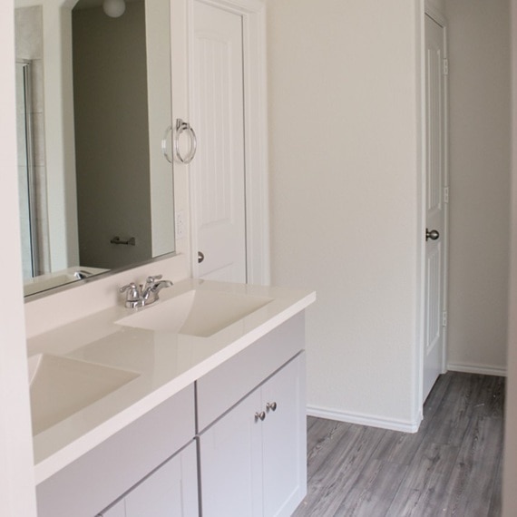 Before photo of bathroom vanity with builder-grade mirror, countertop with dual sinks, sliver faucets, and gray lower cabinets.