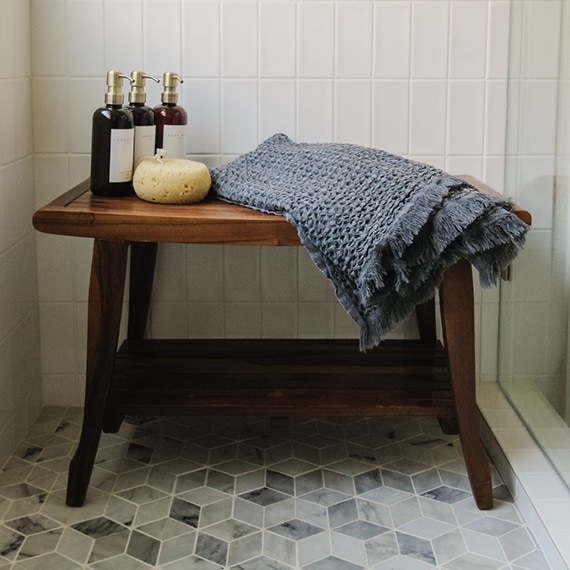 Wood bench holding gray towel, loofah, and haircare products, in shower with gray and white marble mosaic shower floor, and white shower wall tile.