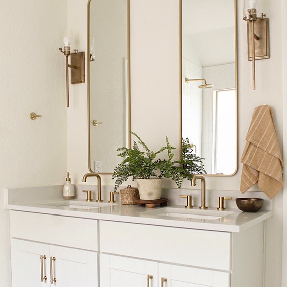 Bathroom vanity with white quartz countertop, white lower cabinets, dual undermount sinks, brass faucets, mirrors with brass frames, and brass wall sconces.