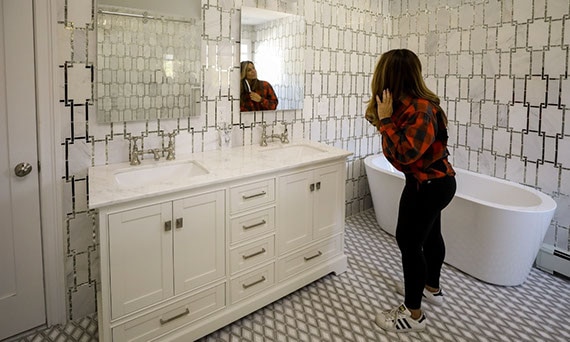 Dolores Catania in her bathroom with white marble & antique mirror wall tile, white marble vanity countertop, white & gray diamond-shaped mosaic floor tile, and soaker tub.