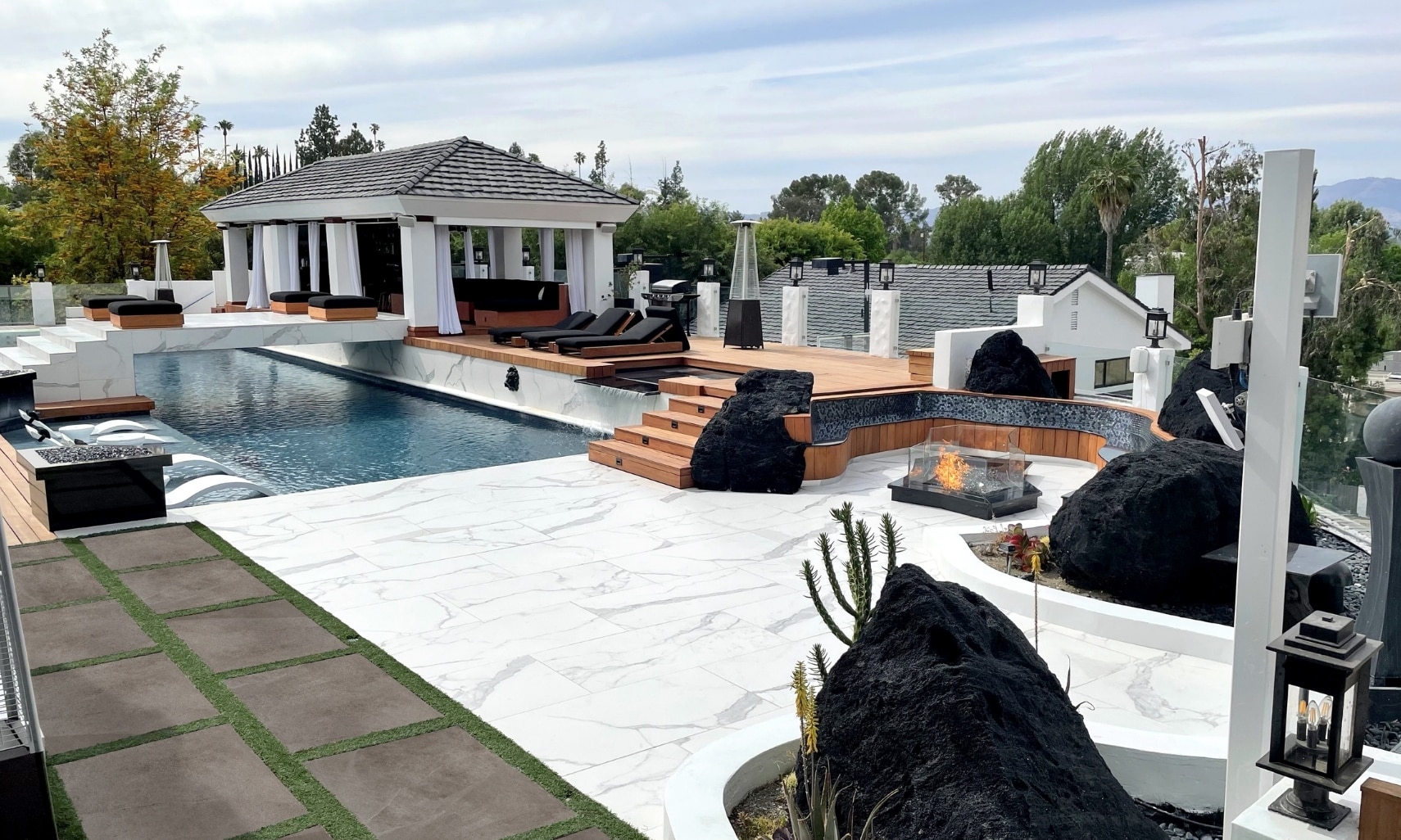 Jason Derulo’s backyard with pool deck & bridge made of white & gray porcelain tile that looks like marble, and wood deck with lounge chairs.