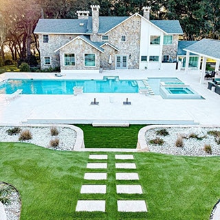View of backyard of two-story stone house with pool with white paver deck, hot tub, firepit, covered patio, and white porcelain pavers set in grass.