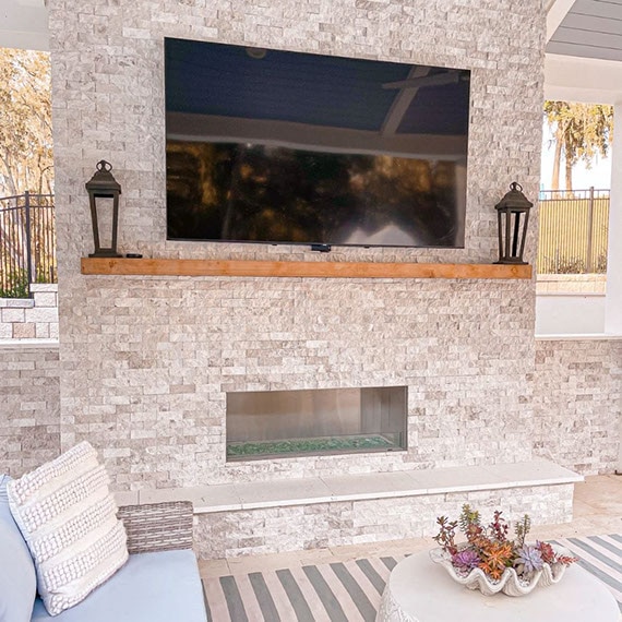 Outdoor fireplace with beige stacked limestone, natural wood mantel holding oil lanterns, wall-mounted big screen TV, gray and white wood patio roof.