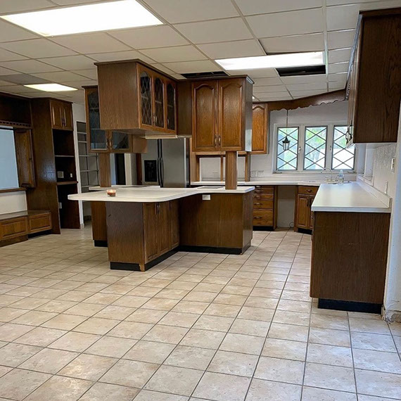 Before photo of kitchen with beige floor tile, white Formica countertops, dark wood upper and lower cabinets, and ceiling tiles with florescent lights.