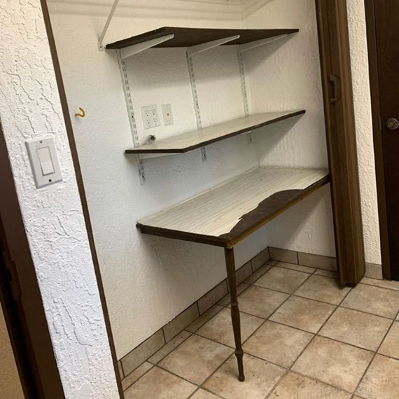 Before photo of mudroom with heavily textured white wall, brown floor tile, damaged shelving, and dark wood door frames.