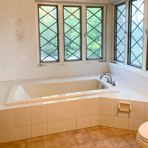 Before photo of bathtub with white tile surround, white wall tile, brown floor tile, and windows with diamond patterns.