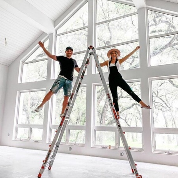 Mark & Laura Beverlin on an A-frame ladder in an unfinished living room with floor-to-ceiling windows.