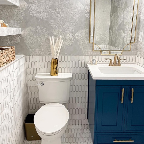 Newly renovated half bath with white & gray marble picket wainscot and floor tile, gray wallpaper with white feathers, blue vanity with brass faucet & accents.