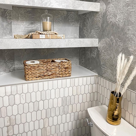 Newly renovated half bath with white & gray marble picket wainscot, gray wallpaper with white feathers, wicker basket & candle on shelves.