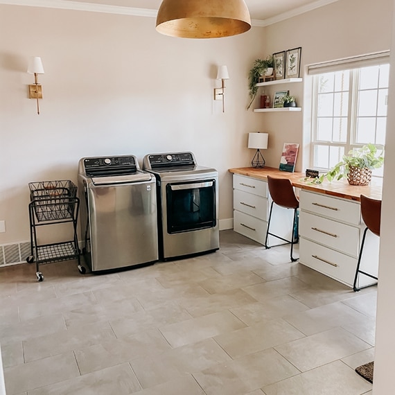 Recently renovated laundry room with gray floor tile that looks like concrete, stainless steel washer & dryer, white cabinets with natural wood countertop.