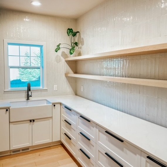 Recently renovated walk-in pantry with white high-gloss wall tile, floating shelves, white quartz countertops, and farmhouse sink in front of window.