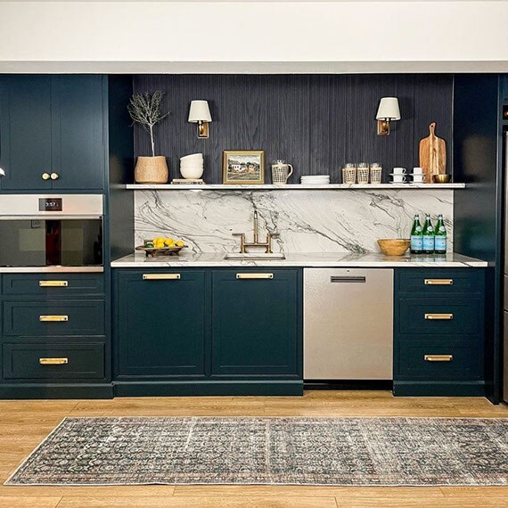 Basement wet bar with dark teal cabinets, brass accents, gray natural quartzite countertop & backsplash, and sink with brass faucet.
