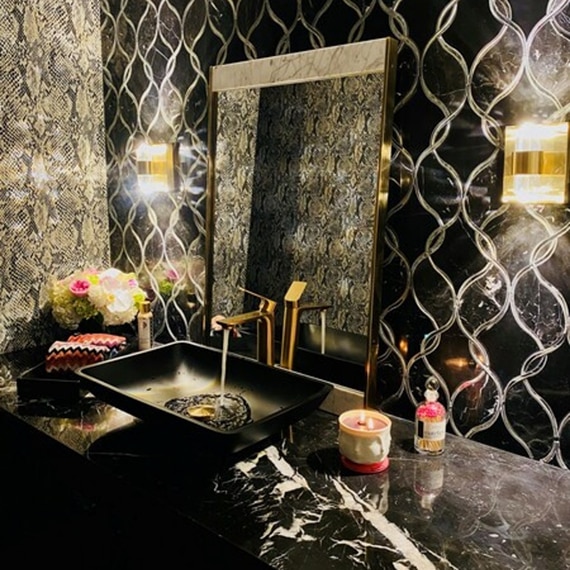 Powder room with black tile with antique mirror accent backsplash, black marble countertop, black vessel sink with brass faucet, and snakeskin wallpaper.