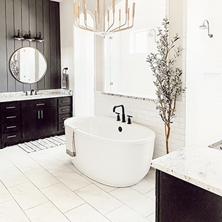 Newly renovated bathroom with white & gray floor tile that looks like marble, soaker tub with black faucet and white backsplash, black vanity with white countertop.