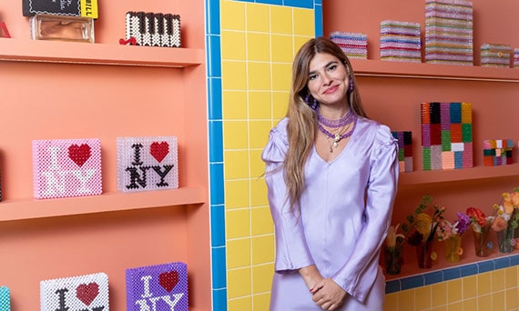 Susan Alexandra in her New York store in front of her accessories on shelves on bright yellow wall tile with bright blue trim tile.