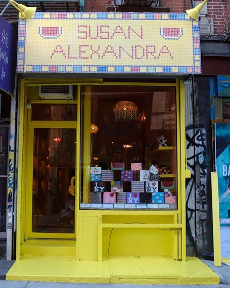 Storefront with neon yellow door and window frame, “Susan Alexandra” sign made of yellow, red, and blue mosaic tile over the door & window.