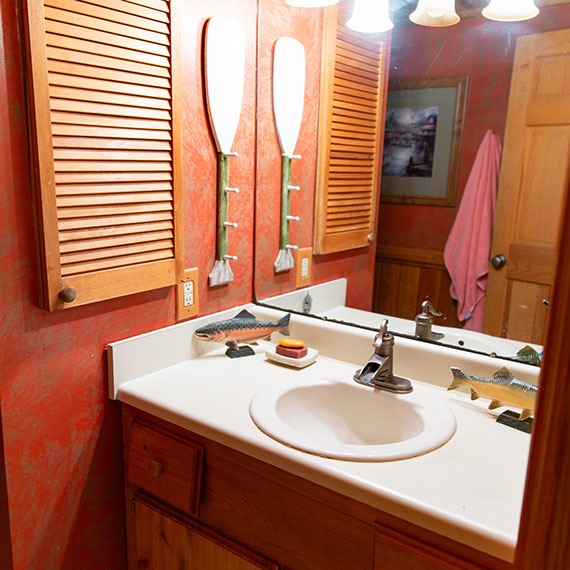Before photo of bathroom with white countertop & sink, wall mirror, red wallpaper, natural wood medicine cabinet and vanity.