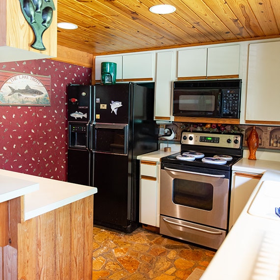 Before photo of kitchen with black refrigerator, white cabinets, red wallpaper, stone flooring, and natural wood ceiling.