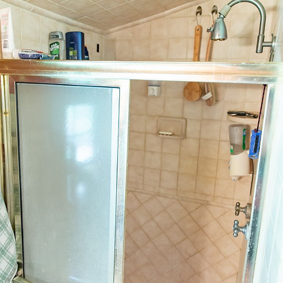 Before photo of bathroom with beige tile shower wall, frosted glass sliding door in silver frame and silver polished showerhead.