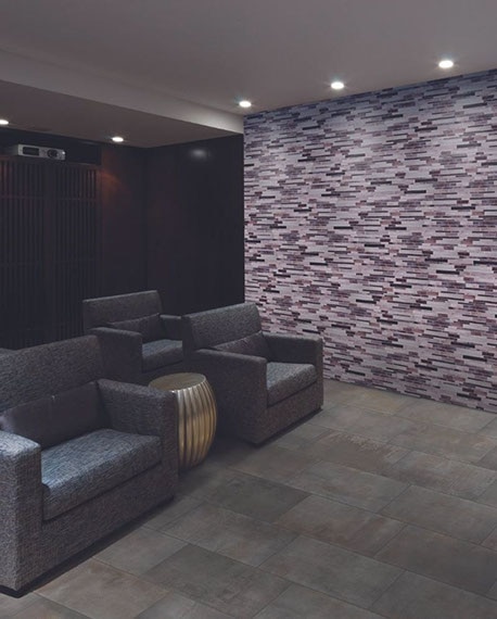 Media room with purple & lilac glass mosaic feature wall, mounted projector, projector screen, gray stone look floor tile, and 4 gray recliners.
