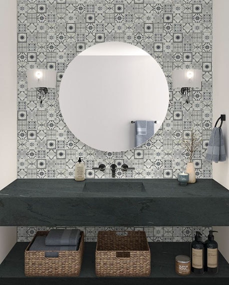 Bathroom vanity with black soapstone countertop and floating shelf, white & black encaustic tile backsplash, wall sconces on each side of round mirror.