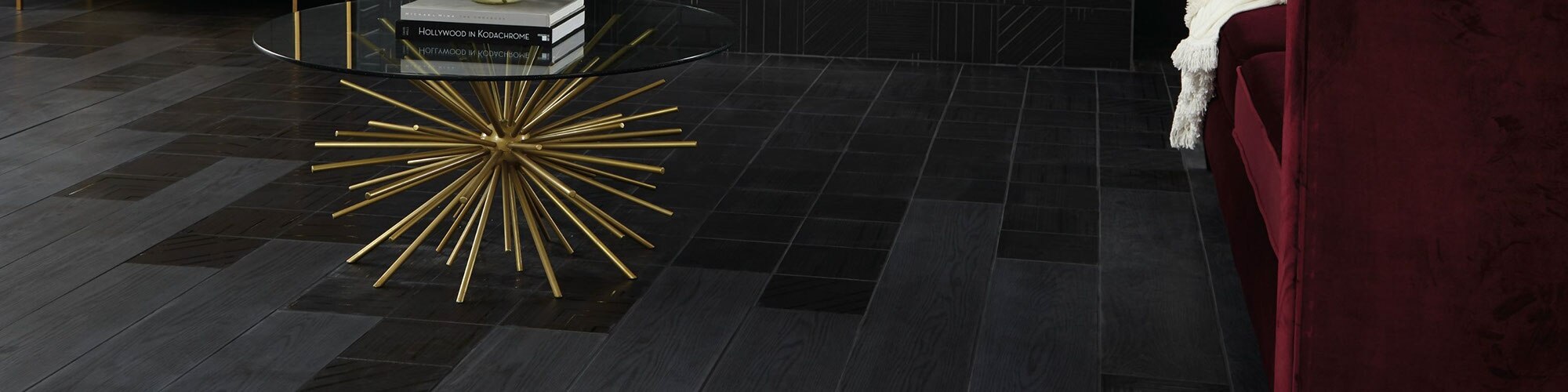Closeup of black wood look tile accented by black geometric tile, crimson velvet chairs, and glass coffee table with gold base.