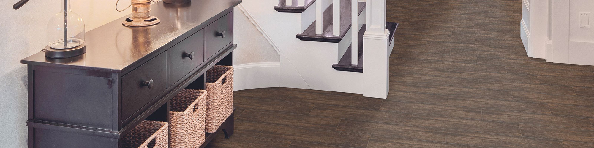 Residential foyer with floor tile that looks like dark wood flooring, dark wood entry table with wicker baskets, staircase with dark wood step & white railing.