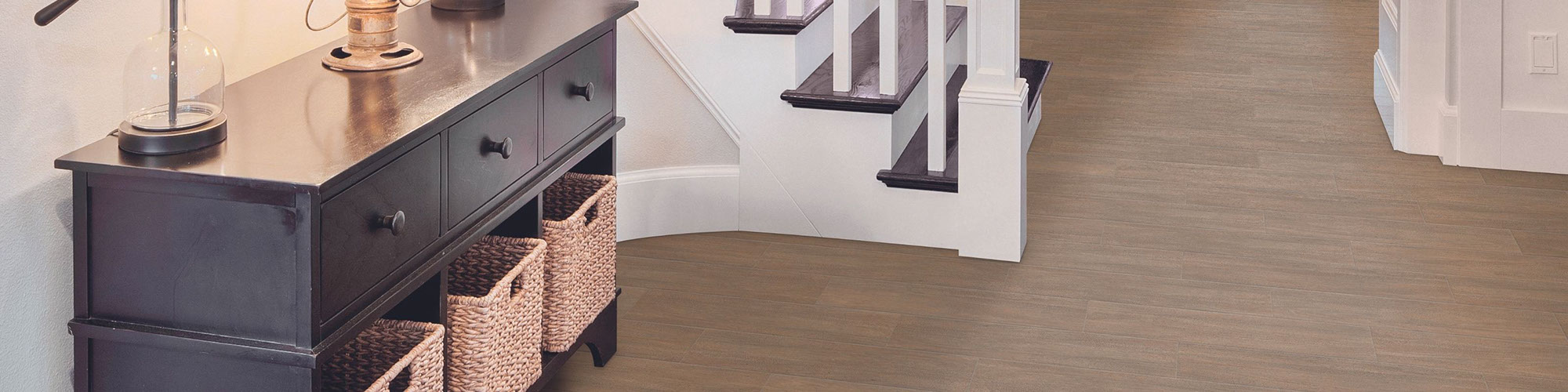 Residential foyer with floor tile that looks like wood flooring, dark wood entry table with wicker baskets, staircase with dark wood step & white railing.