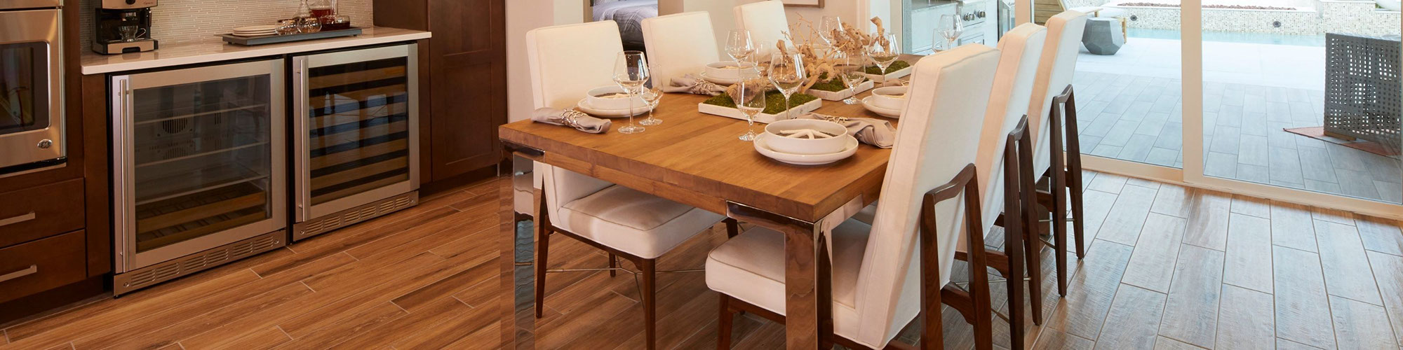 Dining room with floor tile that looks like wood flooring, wood table and white cushioned chairs, white place settings, stemmed wine & water glasses.