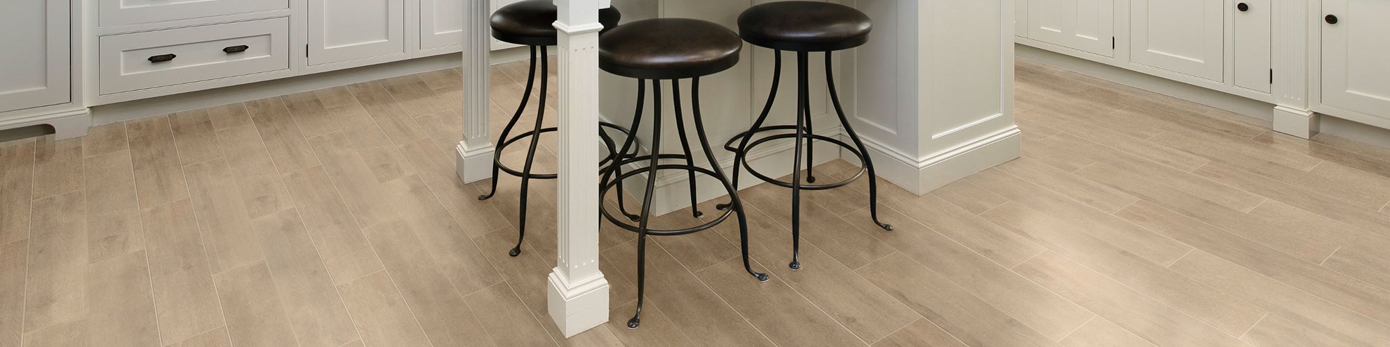 Eat-in kitchen with island with white base, floor tile that looks like blonde wood flooring, brown leather & black metal bar stools.