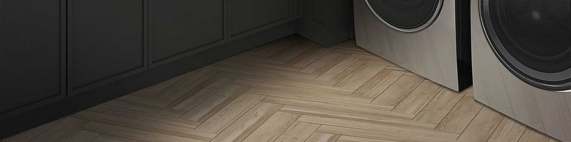 Closeup of laundry room floor of with tile laid in a herringbone pattern that looks like wood flooring, front-loading washer and dryer, and brown cabinets.