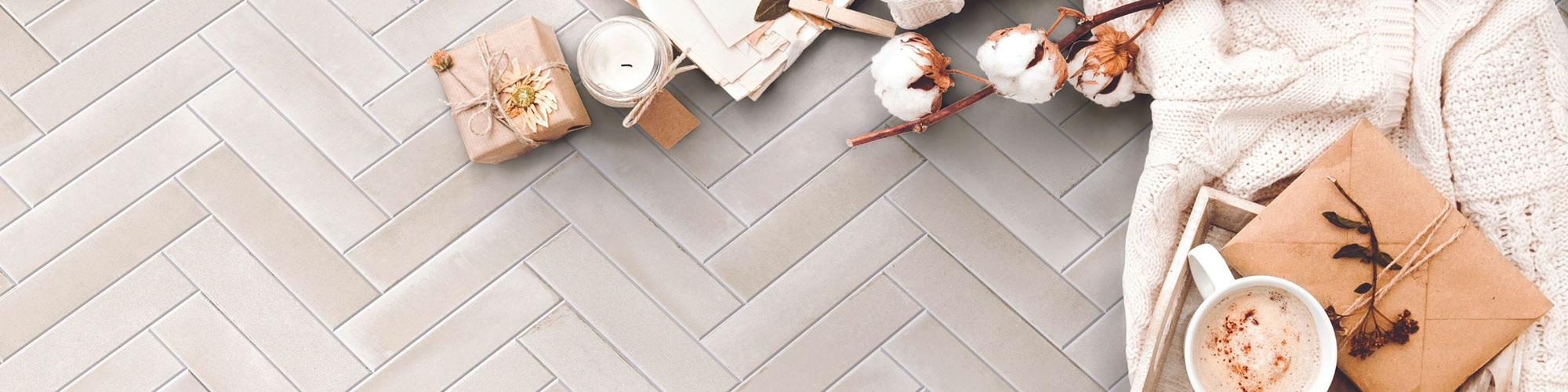 Closeup birds eye view of beige herringbone floor tile that looks like concrete, off-white sweater, latte in mug, stems of cotton bolls and candle wrapped in twine.