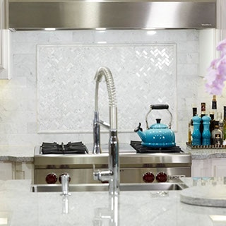 Kitchen with white granite countertop, island with sink, white marble backsplash with raised pencil trim outline, stainless steel stove and vent hood.
