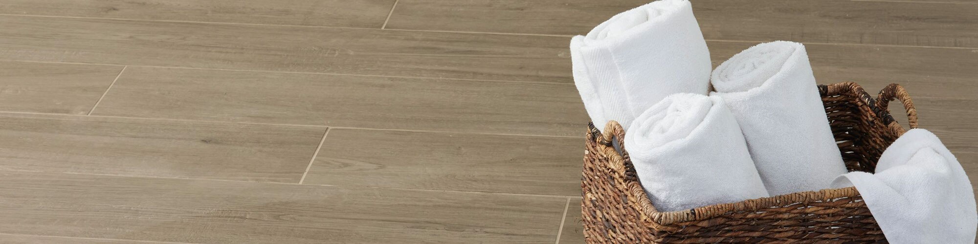 Closeup of wood look tile flooring planks with matching grout, natural wicker basket holding 3 rolled white towels.