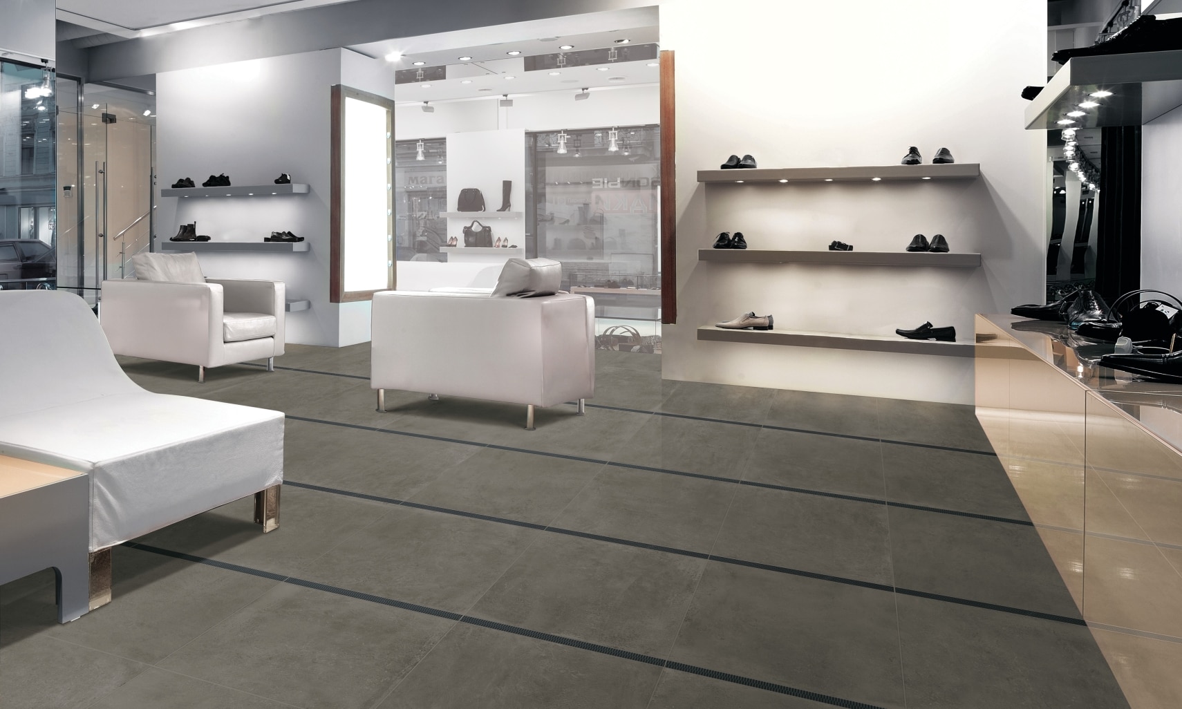 Luxury men’s shoe store with dark gray floor tile that looks like concrete, white leather chairs, and floating shelves holding shoes.