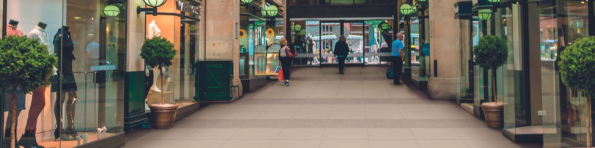 Shopping center with beige & tan floor tile that looks like concrete, glass front boutiques with window dressing and potted plants.